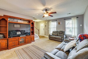 A living room with a large wooden entertainment center, a gray leather sofa, a ceiling fan, and a view of the outside through a window. - Wooded Trail On-site, Spacious Simpsonville Home (Simpsonville)