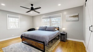 Un dormitorio con una cama, dos mesitas de noche, un ventilador de techo y dos ventanas.