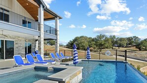 A swimming pool with blue lounge chairs and umbrellas, a stone building with a balcony, and a clear sky. - Waterfront Home on Lake LBJ w/ Pool & Dock! (Kingsland)