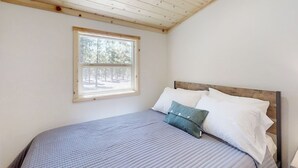 A bedroom with a wooden ceiling, a window with a view of trees, a bed with a blue and white bedspread, and a wooden headboard.