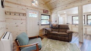 A cabin interior with a wooden ceiling, a cozy living area with a brown sofa, a rocking chair, and a kitchen area with cabinets and a window.