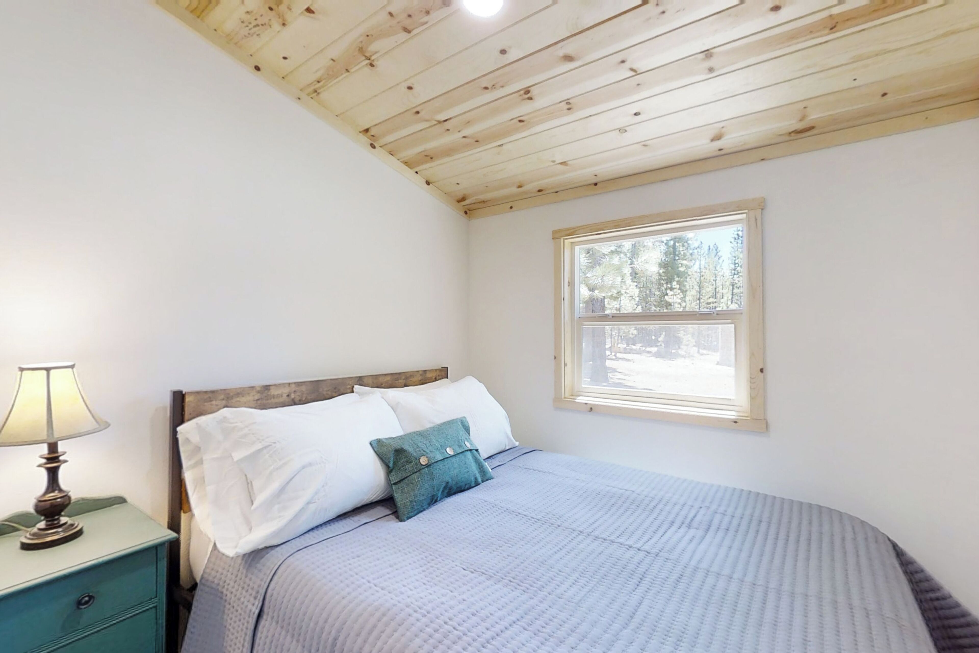 A bedroom with a wooden ceiling, a bed with white and teal pillows, a nightstand with a lamp, and a window with a view of trees.