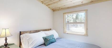 A bedroom with a wooden ceiling, a bed with white and teal pillows, a nightstand with a lamp, and a window with a view of trees.
