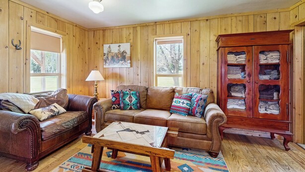 A cozy living room with wooden walls, two leather sofas, a coffee table, and a cabinet with folded blankets. - 1 BR Bunk House on Long Hollow Ranch, Dog Friendly (Sisters)