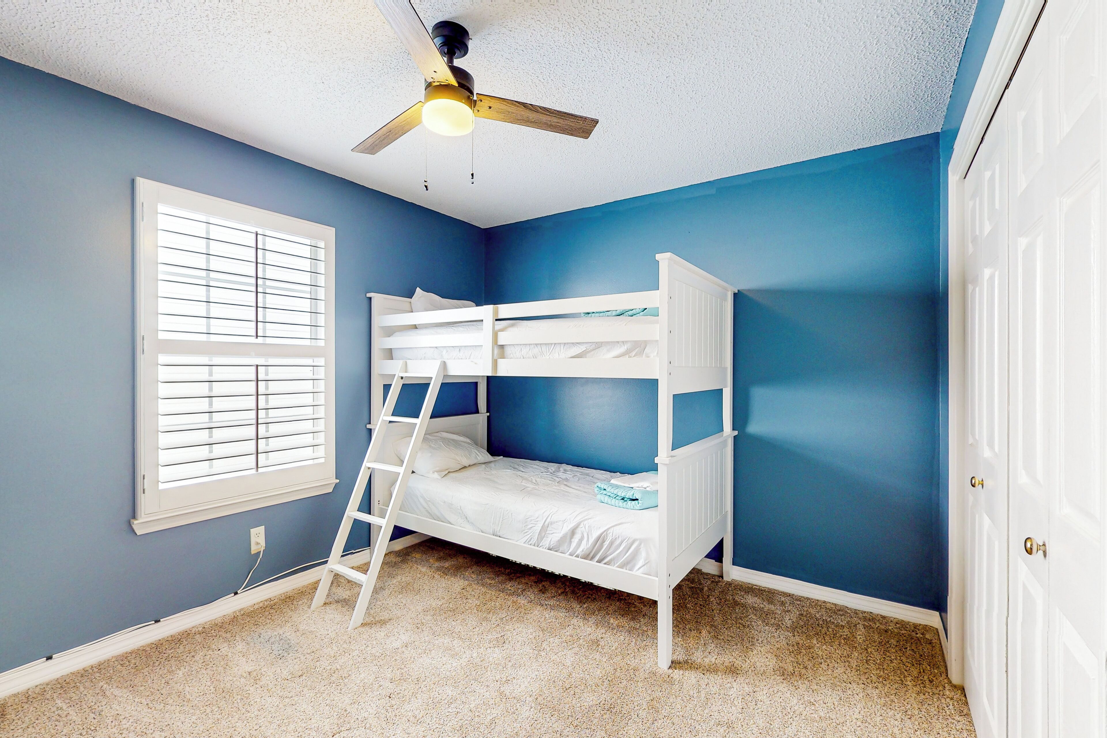 Une chambre avec un lit superposé, un ventilateur de plafond, une fenêtre avec des stores et une porte.