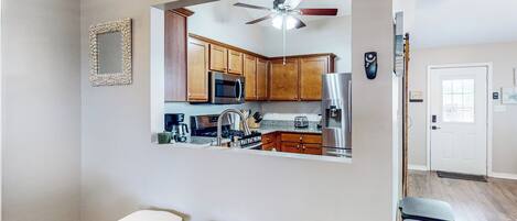 A compact kitchen with wooden cabinets, a microwave, and a ceiling fan. There are three white stools and a small table.