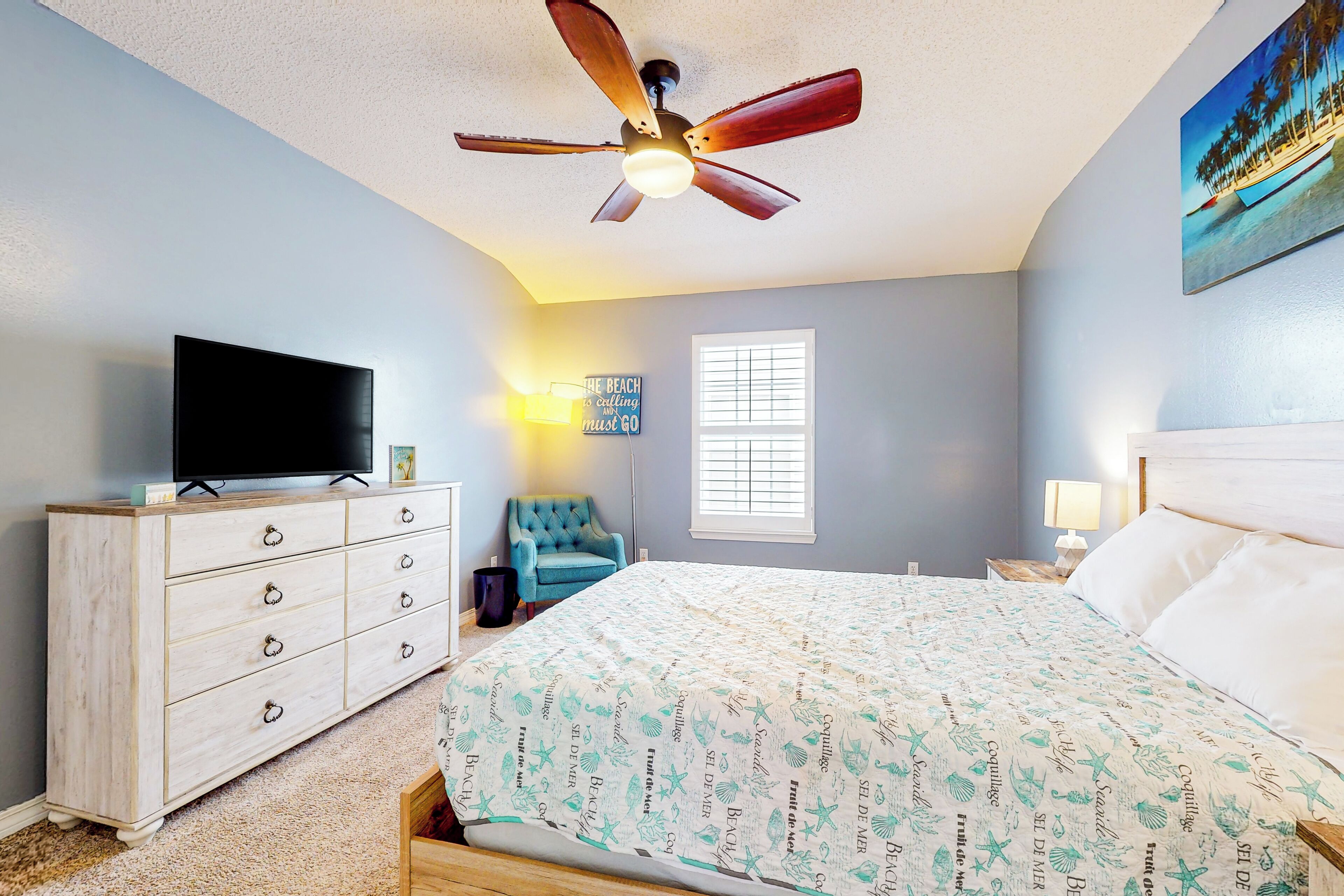 A bedroom with a bed, a TV on a dresser, a ceiling fan, a window, and a painting on the wall.
