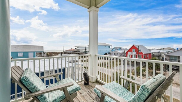 A wooden deck with outdoor furniture, a white railing, and a view of colorful houses in the background. - Fort Morgan beachside retreat ~ pet-friendly ~ close to the sand (Gulf Shores)