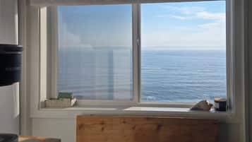 A room with a large window overlooking the sea, a wooden bench, and a small table with a cup.