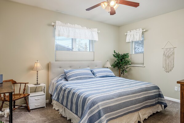 Une chambre à coucher comprenant un lit, des tables de chevet, une chaise, un ventilateur de plafond et une fenêtre avec des rideaux.