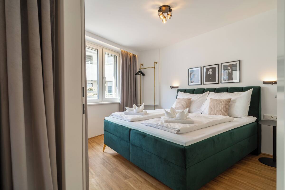Una cama bien hecha con sábanas blancas, un cabecero tapizado en verde y una mesita de noche con una lámpara.