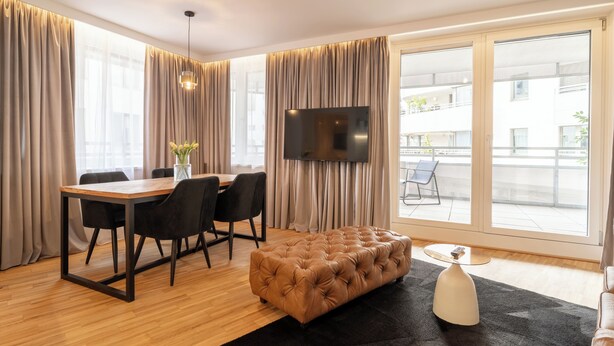 A modern living room with a wooden dining table, black chairs, a tufted ottoman, a flat-screen TV, and large windows with curtains.