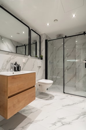 Ein modernes Badezimmer mit Marmorboden, einem Holzkosmetikschrank, einer weißen Toilette und einer Duschkabine aus Glas.