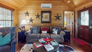 A cozy living room with a wooden ceiling, a brown leather sofa, a coffee table with books, and decorative items. - Water-view Cabin w/ Front Porch, W & D, Streaming (Carmine)
