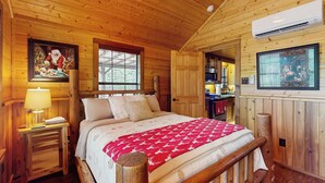 A cozy log cabin bedroom with a wooden bed, a nightstand with a lamp, and a large window. - Water-view Cabin w/ Front Porch, W & D, Streaming (Carmine)