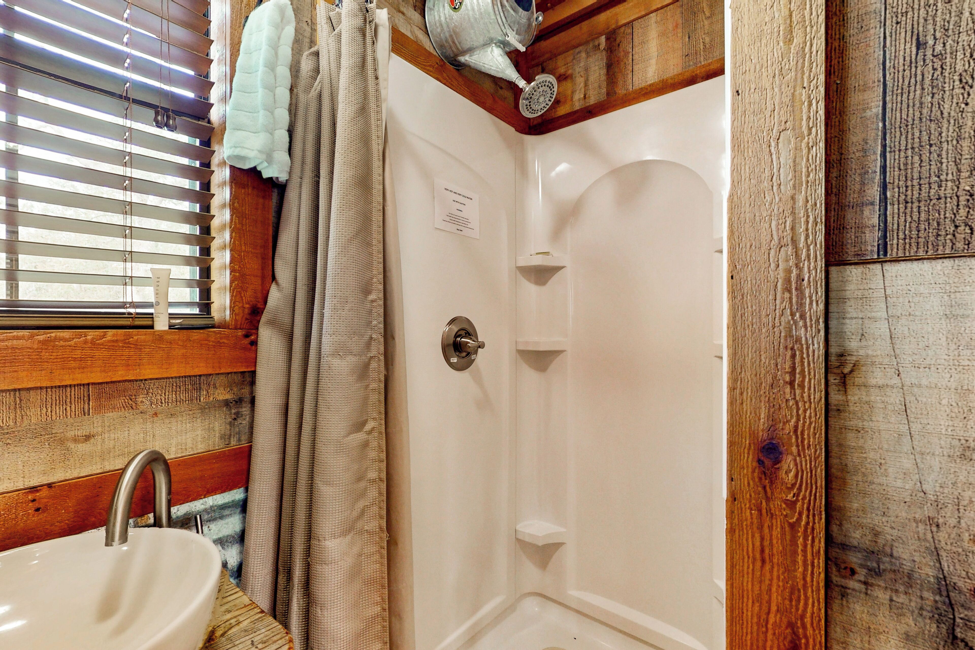 Un baño con suelo de madera, un lavabo blanco, una ducha con cabezal de ducha y una ventana con persianas.