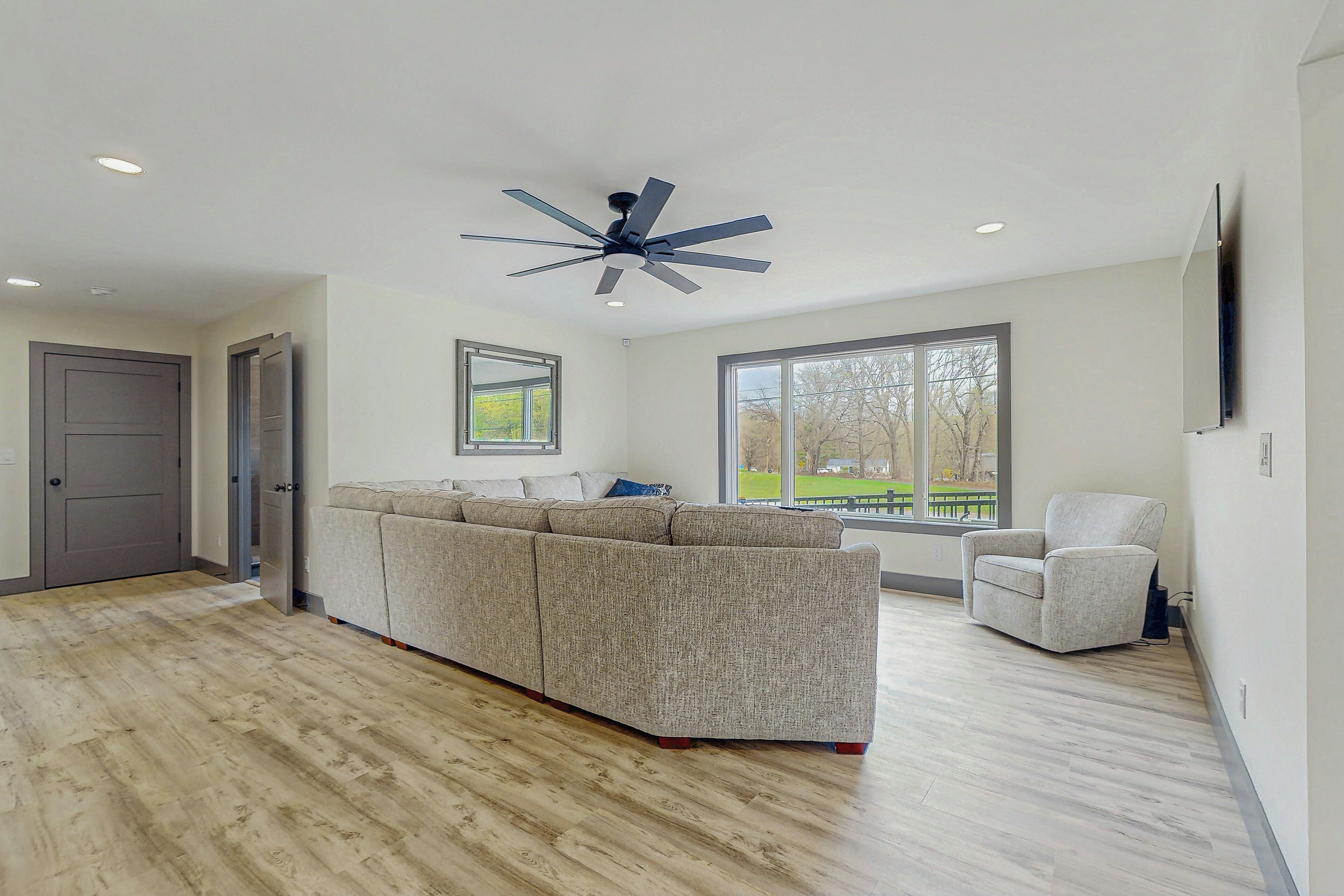 Un salon moderne avec un canapé d’angle, un ventilateur de plafond et une grande fenêtre donnant sur l’extérieur.