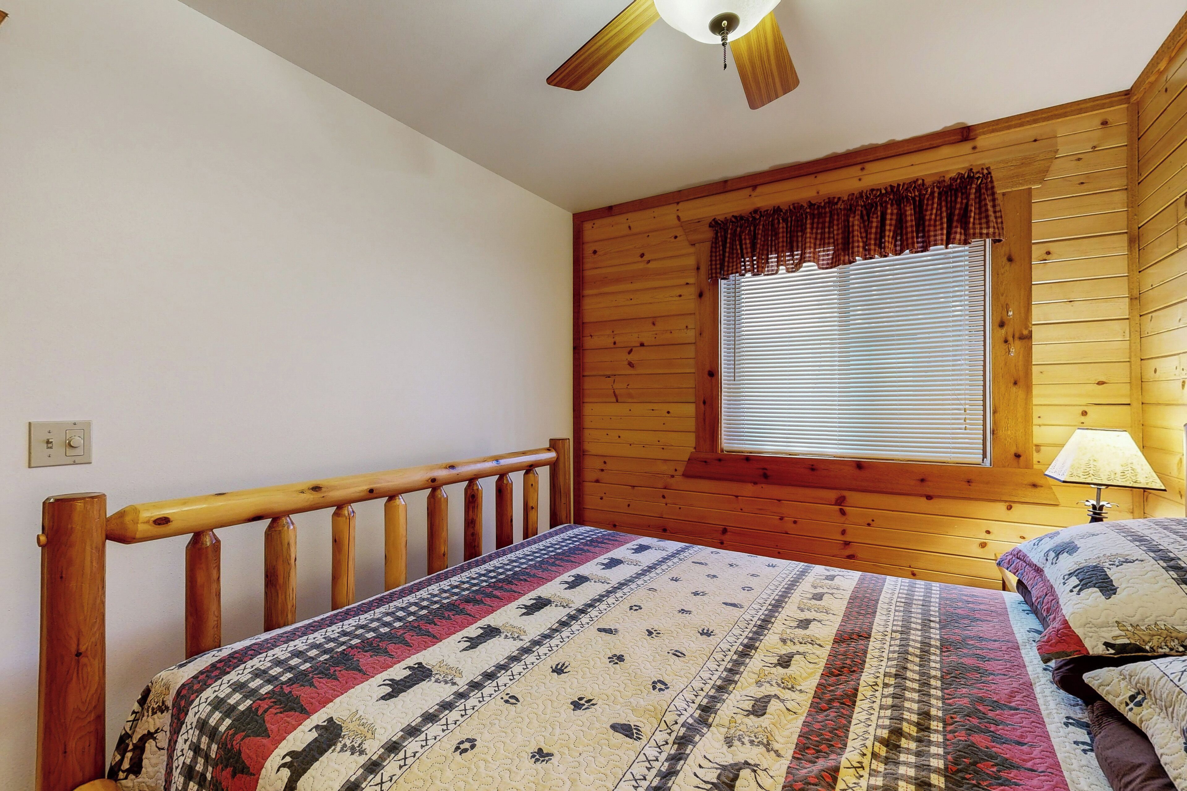 A wooden cabin bedroom with a bed, a window with blinds, a ceiling fan, and a lamp on the nightstand.