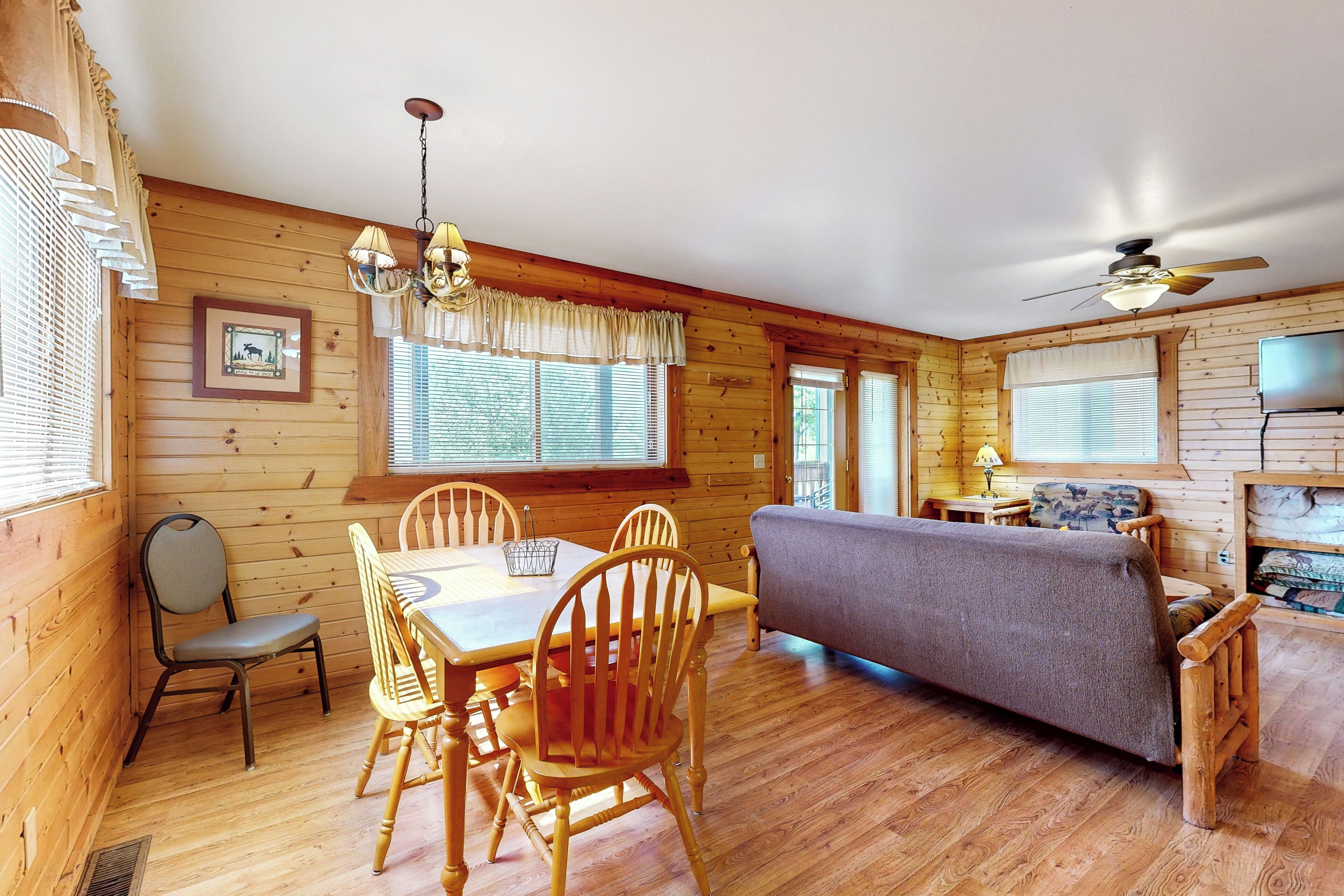 A wooden cabin interior with a dining table, chairs, a sofa, and a ceiling fan.