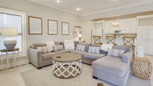 Una sala de estar moderna con un sofá modular, una mesa de centro redonda y una cocina con armarios blancos y una lámpara colgante.