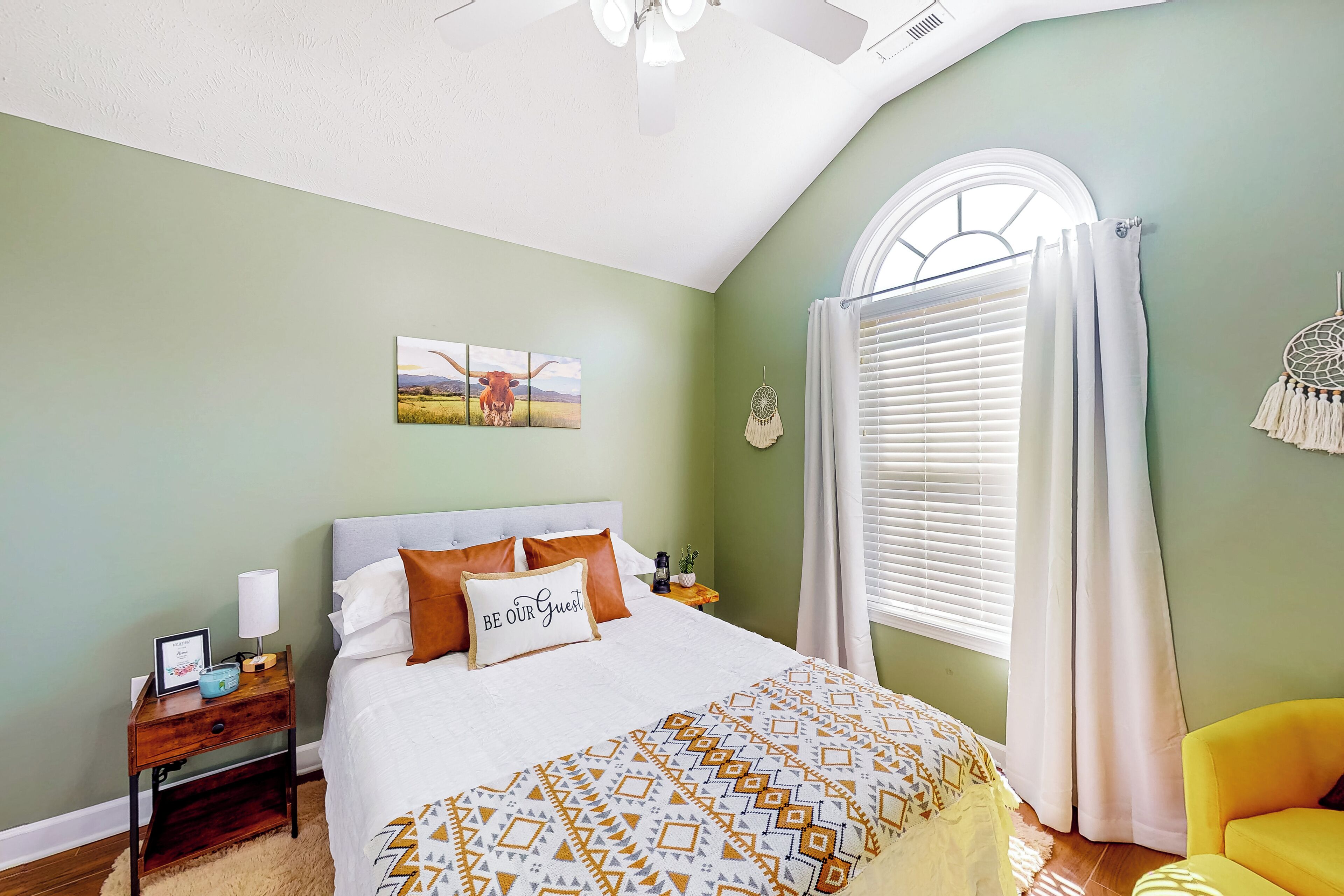 A bedroom with a bed, a nightstand, a window with curtains, a dreamcatcher, and a yellow chair.