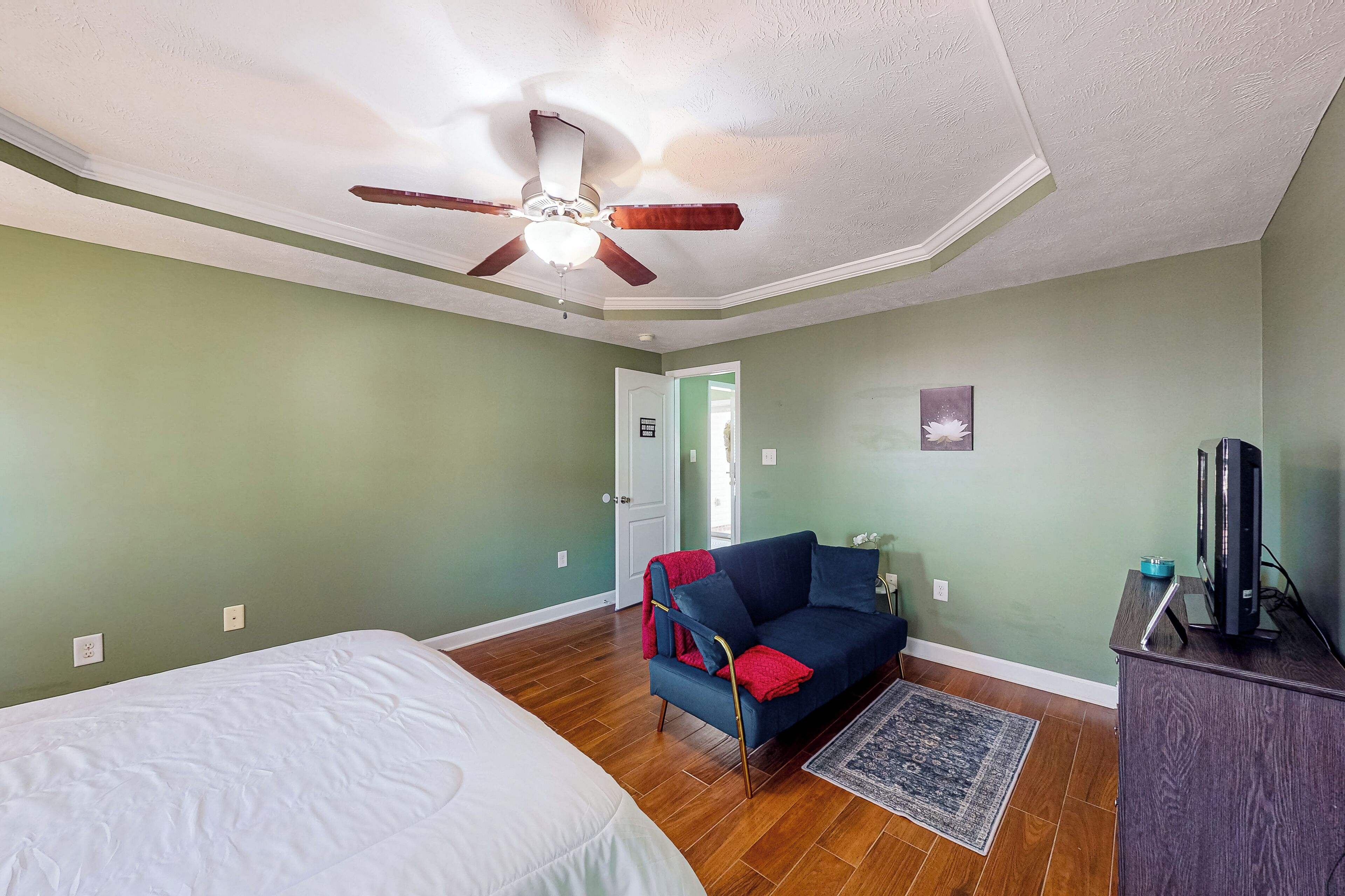 Una habitación con una cama, un sofá azul con cojines rojos, un ventilador de techo, un televisor sobre un mueble bajo y una alfombra.