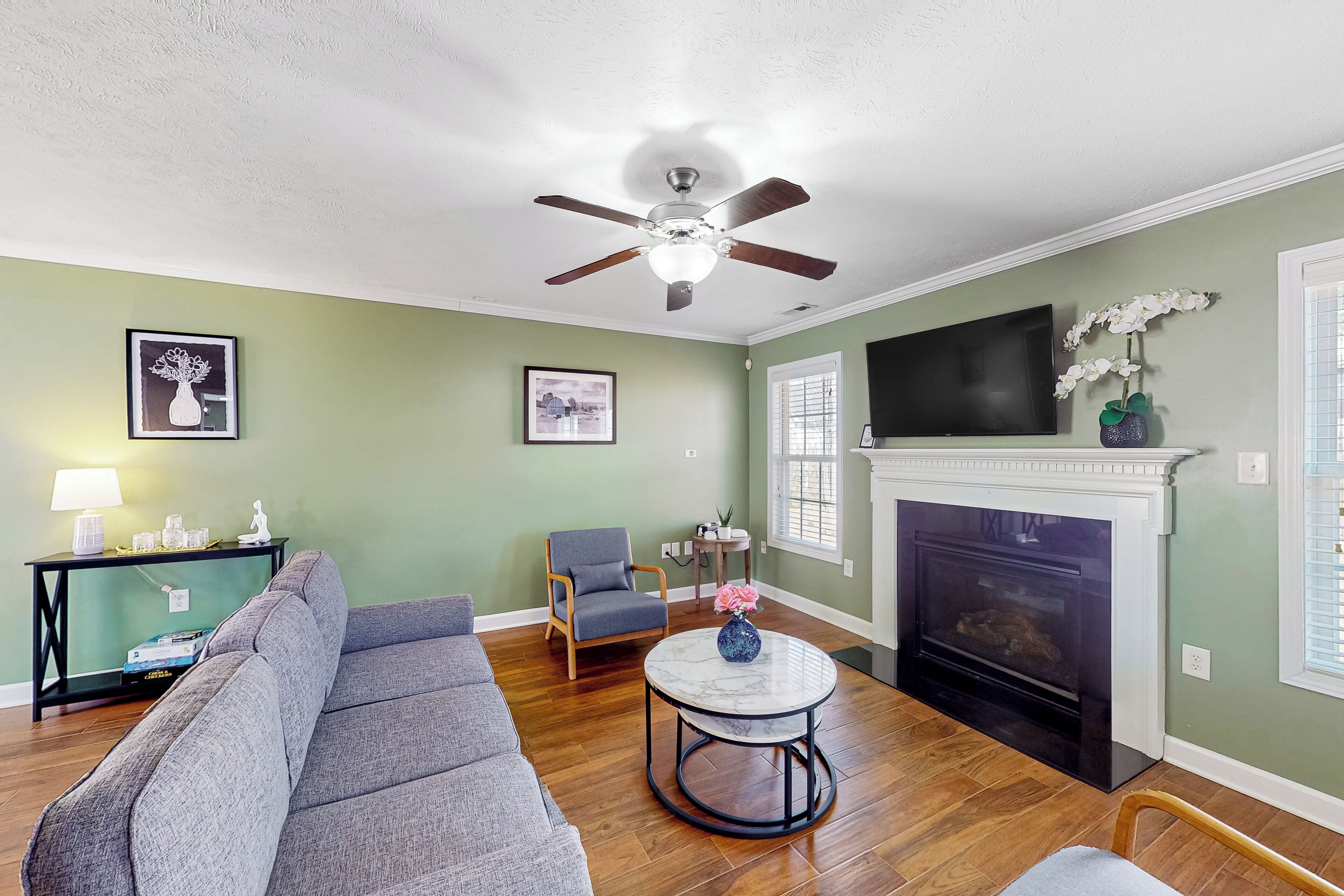 A living room with a gray sofa, a round coffee table, a ceiling fan, a fireplace, and a TV mounted on the wall.