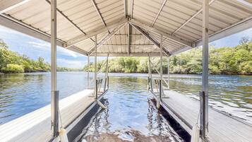 A covered dock with a metal roof and wooden planks, overlooking a calm river with dense greenery in the background.