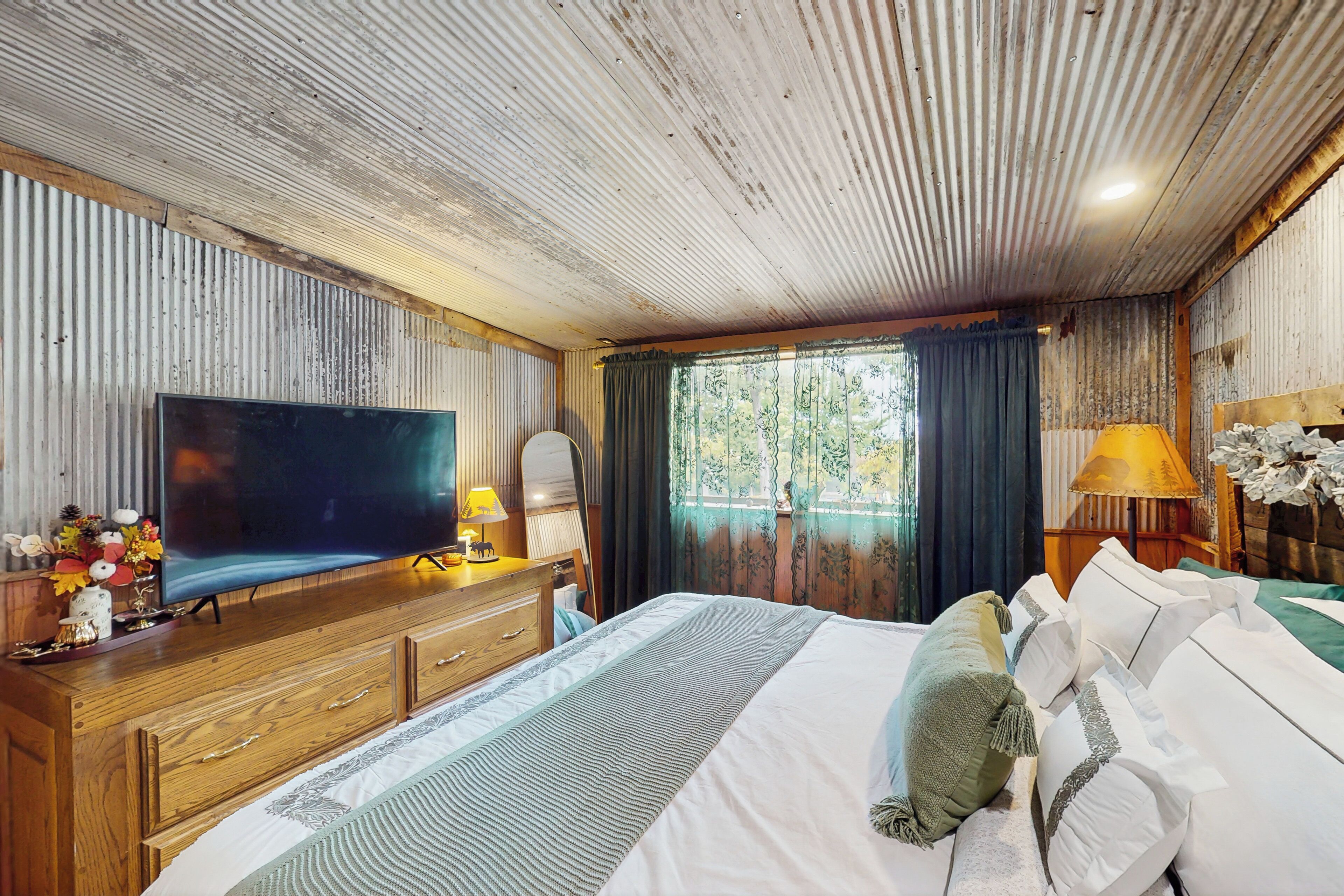 Ein Schlafzimmer mit Holzboden, einem großen Flachbildfernseher, einem Bett mit weißer Bettwäsche, einer hölzernen Kommode und einem Fenster mit Vorhängen.