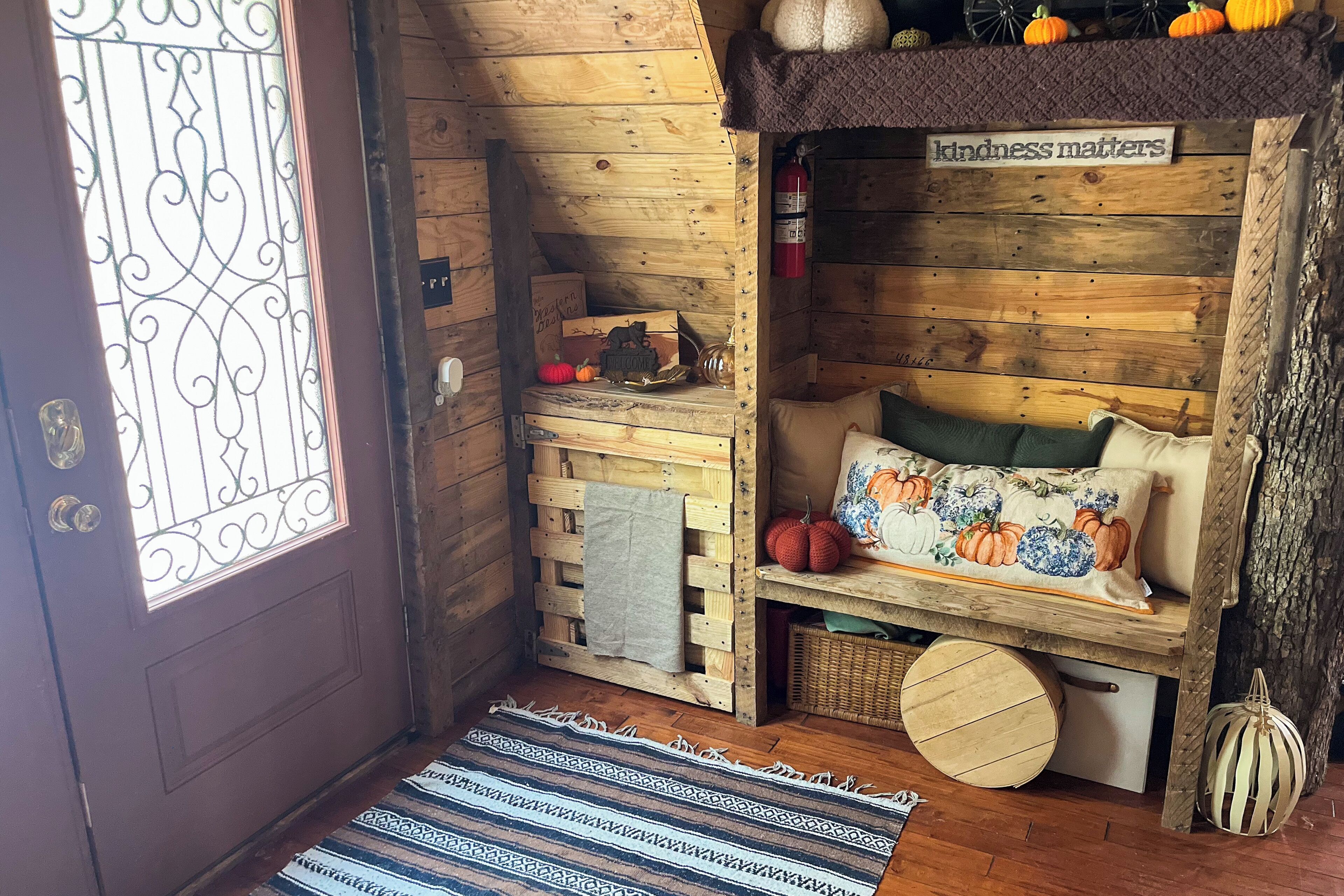 L’intérieur chaleureux d’un chalet avec une porte en bois, un banc agrémenté de coussins décoratifs, une étagère avec des citrouilles et un panneau indiquant : « La gentillesse est importante ».