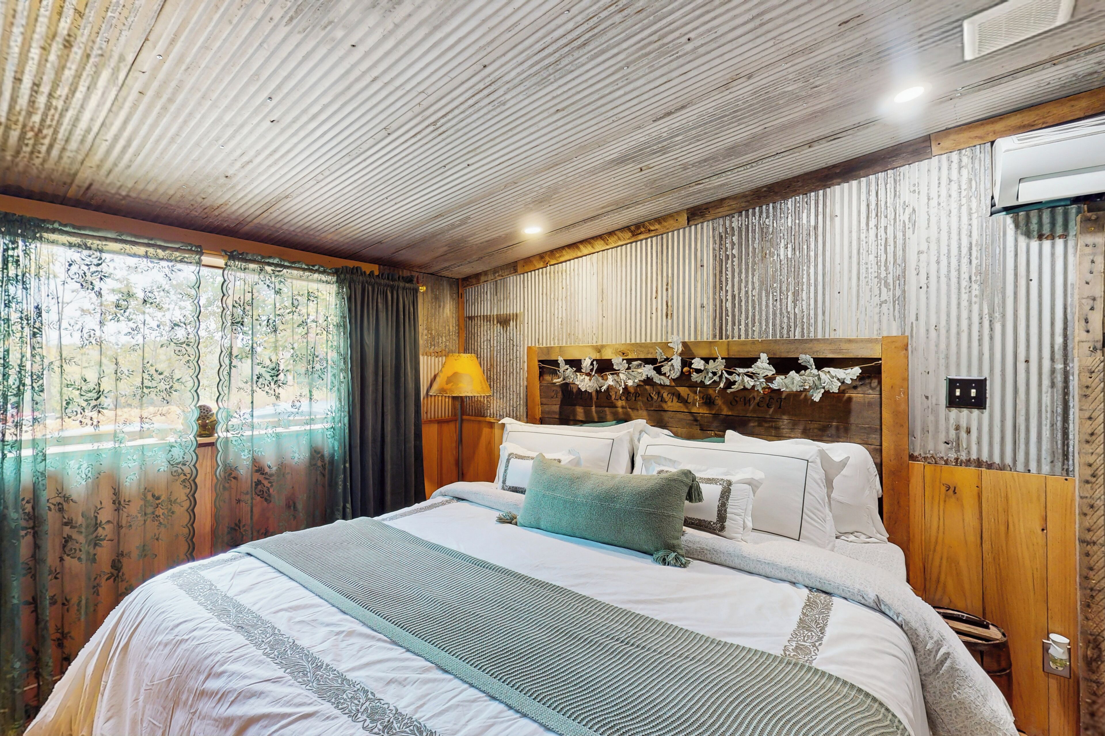 Un dormitorio con cabecera de madera, una cama grande con varias almohadas, una mesita de noche con lámpara y vistas a través de una ventana con cortinas.