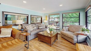 A spacious living room with a large window, a sofa, a coffee table, and a bookshelf. - 2 BR Lakefront House w/ Hot Tub in ADK Park (Gloversville)