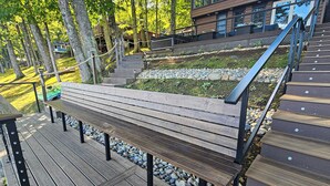 A wooden deck with a bench, surrounded by trees and a stone pathway. - 2 BR Lakefront House w/ Hot Tub in ADK Park (Gloversville)