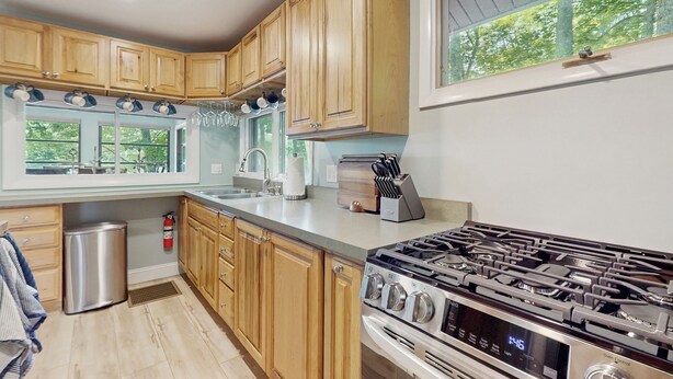 A kitchen with wooden cabinets, a stainless steel range, and a window with a view of trees. - 2 BR Lakefront House w/ Hot Tub in ADK Park (Gloversville)