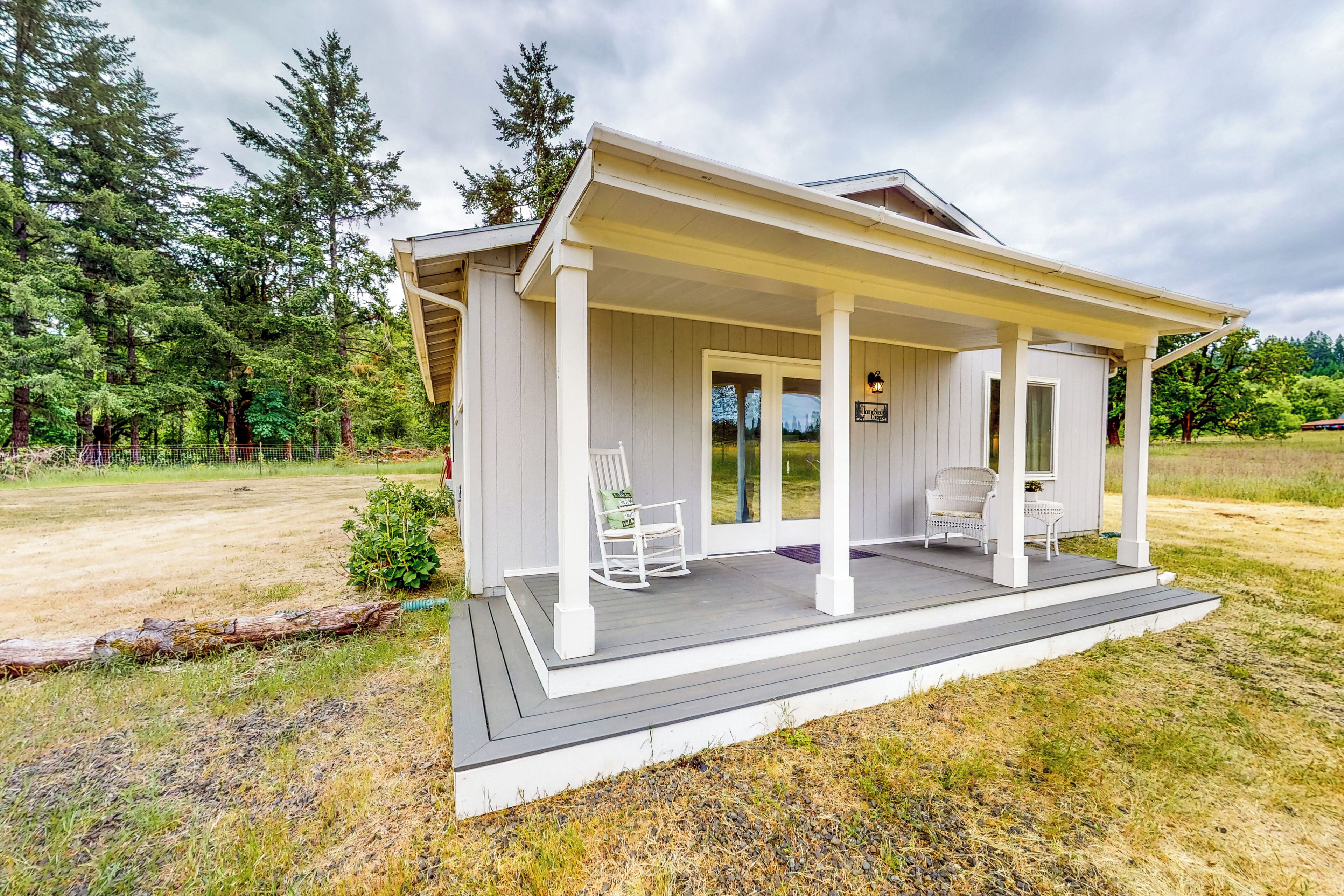 A single-story house with a covered porch, a small porch area with two chairs, and a view of the surrounding forest.