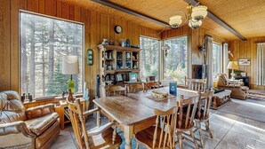 A wooden-paneled dining room with a large table and chairs, a fireplace, and a view of the forest through large windows. - 3BR Rustic Riverfront Home w/ Wood Stove (Blairsden-Graeagle)