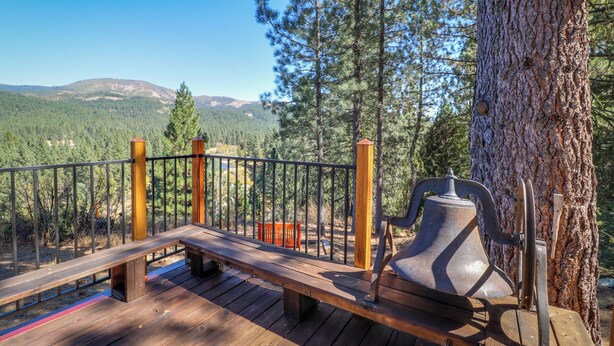 A wooden deck with a bench and a bell, overlooking a forested area with mountains in the background. - 3BR Rustic Riverfront Home w/ Wood Stove (Blairsden-Graeagle)