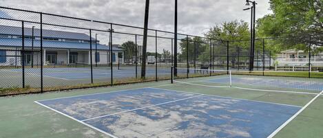 A tennis court with a net, surrounded by a chain-link fence, and a building with a blue roof in the background.