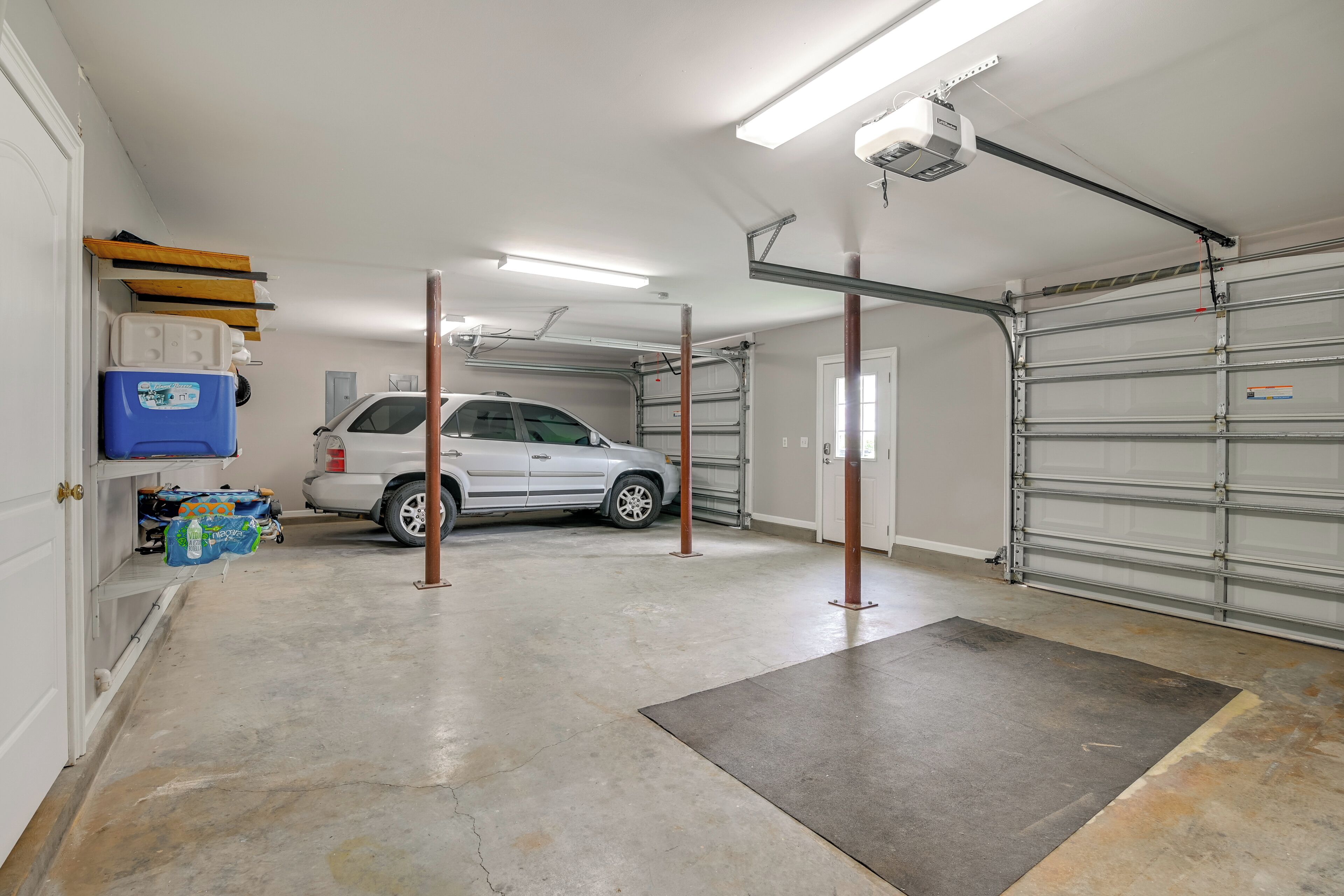 A garage with a white SUV parked inside, a blue cooler on the left, and a garage door on the right.
