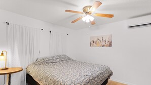 Un dormitorio con cama, ventilador de techo, aire acondicionado de pared y un cuadro.