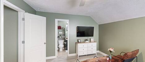 A room with a ceiling fan, a TV mounted on the wall, a small table with a vase, and a cushioned bench.