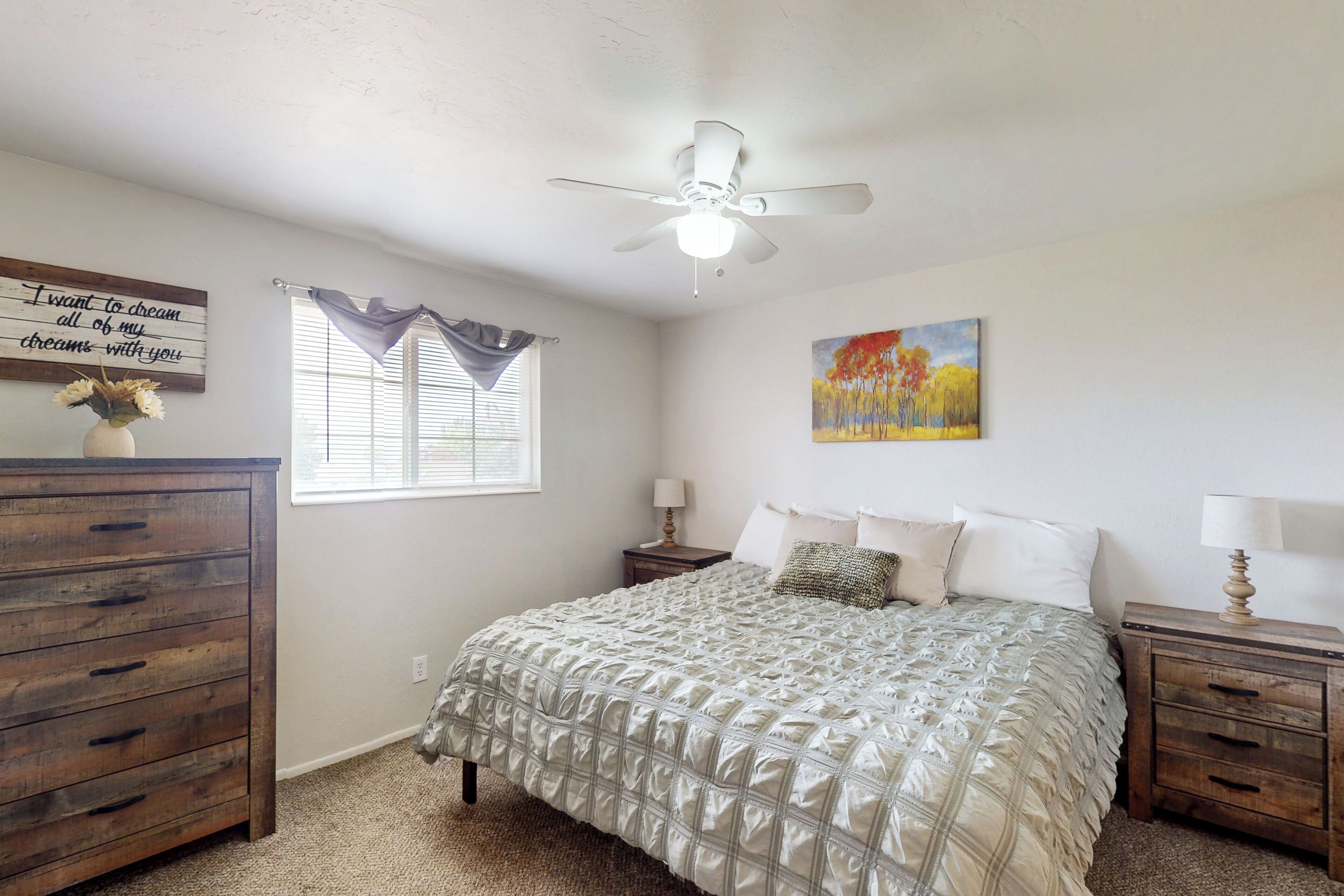 A bedroom with a bed, wooden dresser, nightstands, a ceiling fan, a window with blinds, and a painting on the wall.