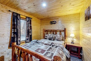 A wooden cabin room with a bed, curtains, a nightstand, and a lamp. - Hot Tub & Fire Pit: Wooded Indiana Escape (Columbus)