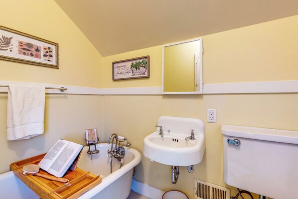 A bathroom with a bathtub, sink, and toilet. There is a towel rack with a towel, a framed picture on the wall, and a wooden tray with a book and brush. - Forbestown Retreat: Easy Access to Clear Lake (Lakeport)