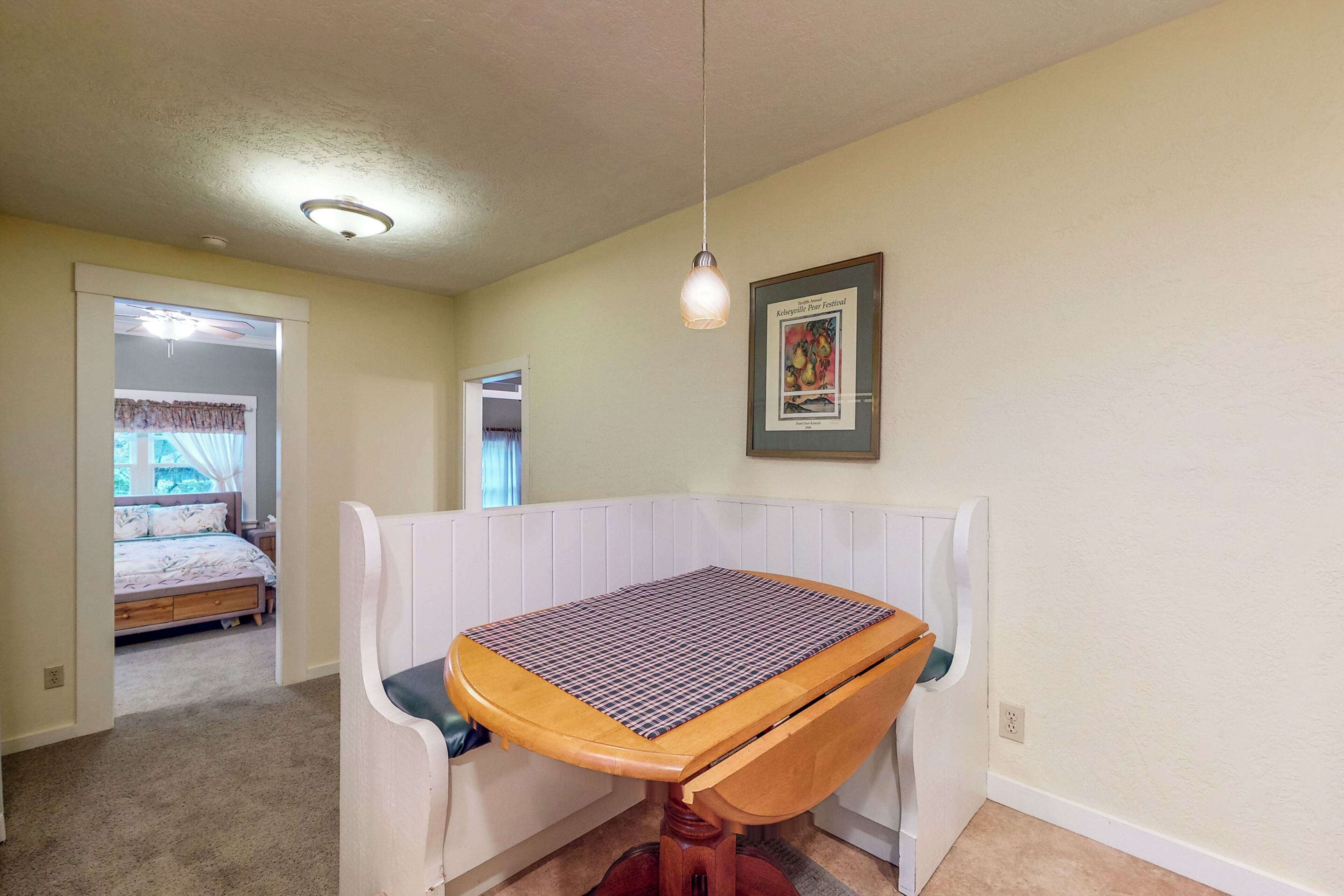 A room with a wooden table and bench, a ceiling light, a framed picture on the wall, and a doorway leading to another room with a bed.