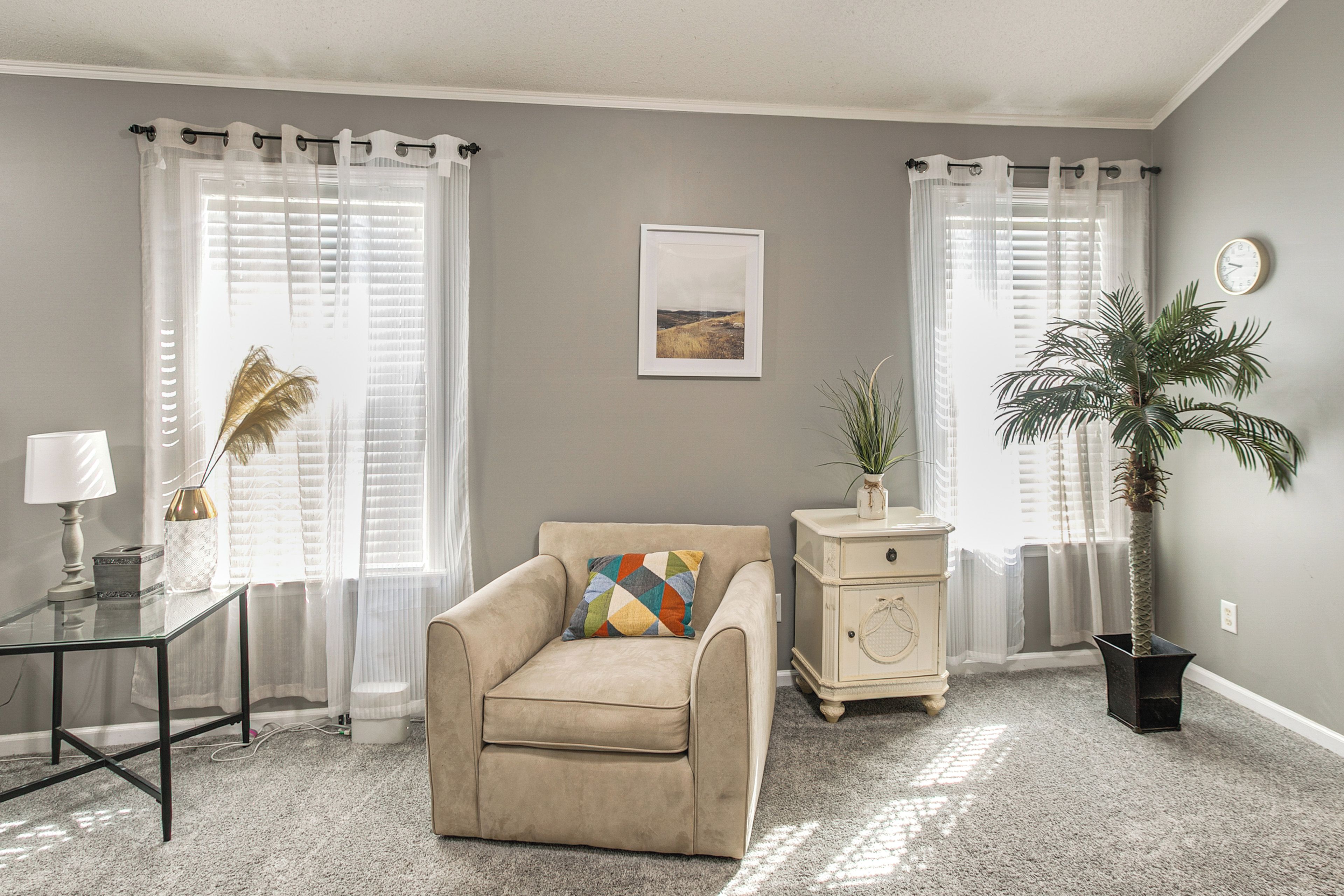A living room with a beige armchair, a small table with a lamp, a decorative plant, and a vintage-style cabinet.