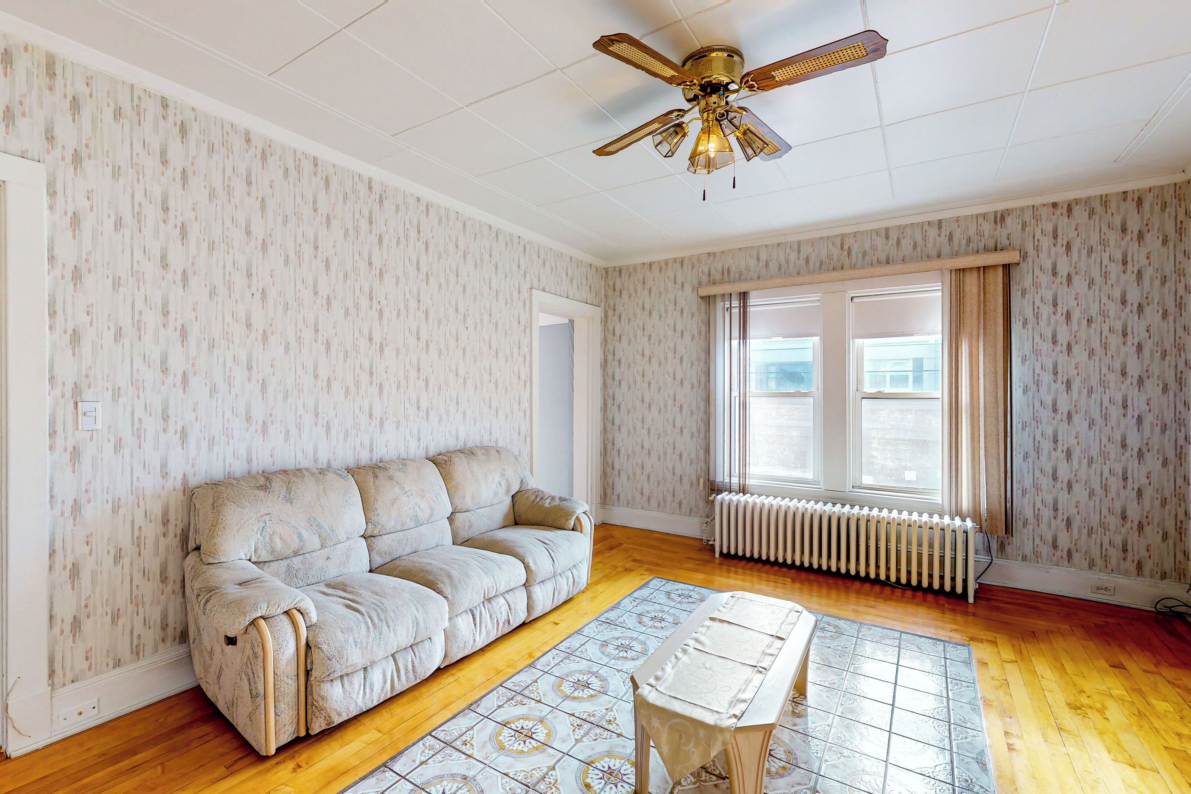 A room with a beige sofa, a patterned rug, a small table, a ceiling fan, and a window with curtains.