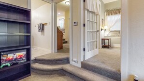 Un couloir avec un escalier recouvert de moquette menant à une pièce dotée d’une porte, d’une étagère sur laquelle se trouve une console de jeux vidéo, et d’une petite table avec une lampe.