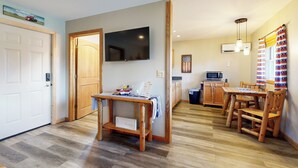 A spacious living area with a wooden dining table and chairs, a flat-screen TV mounted on the wall, and a kitchen in the background.