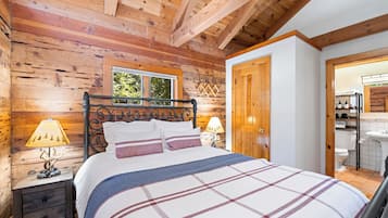 A wooden cabin bedroom with a bed, two bedside lamps, a window, and a door leading to a bathroom.
